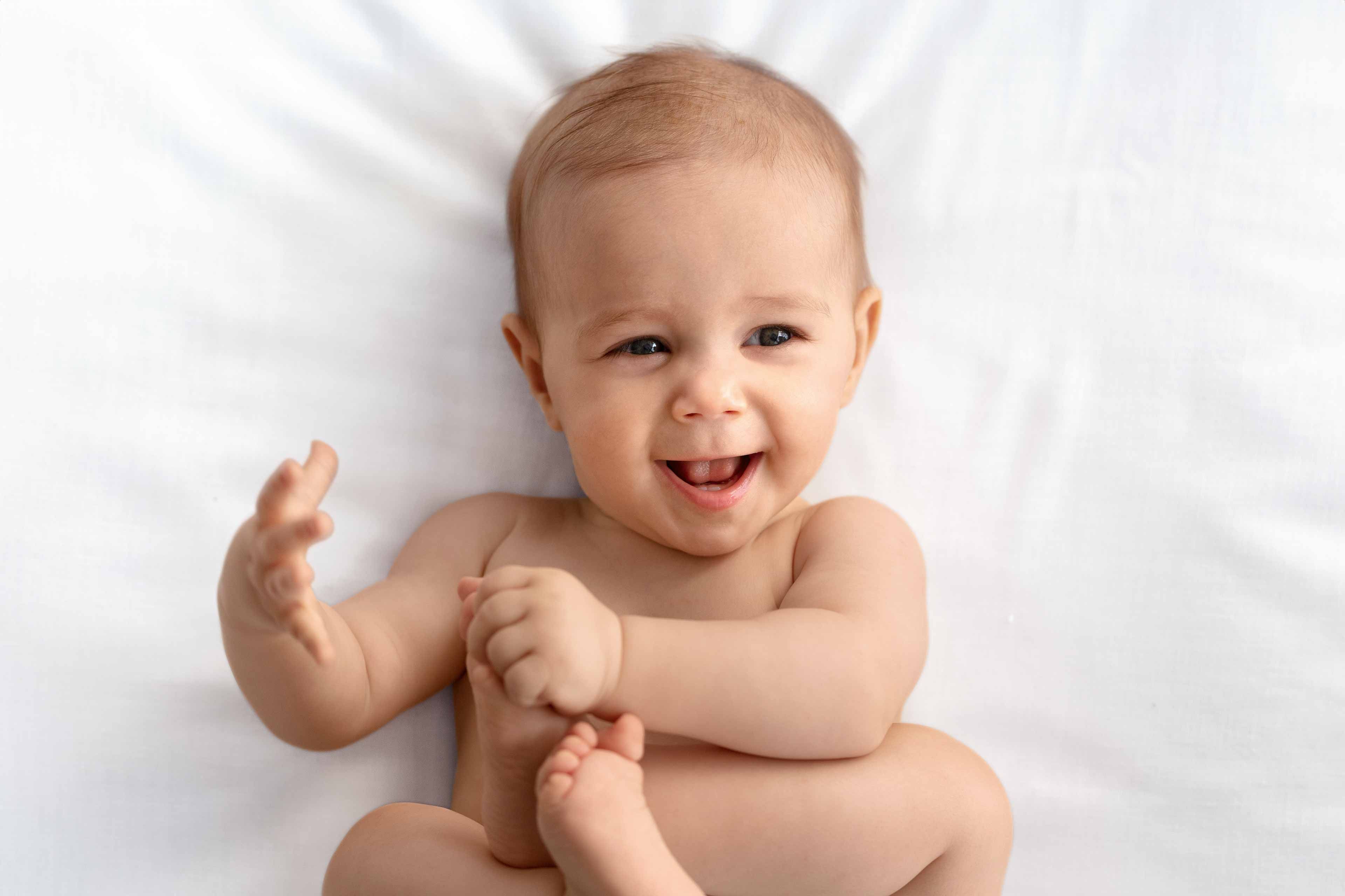 Ein Baby liegt auf einer weißen Fläche, lächelt und spielt mit seinen Füßen.