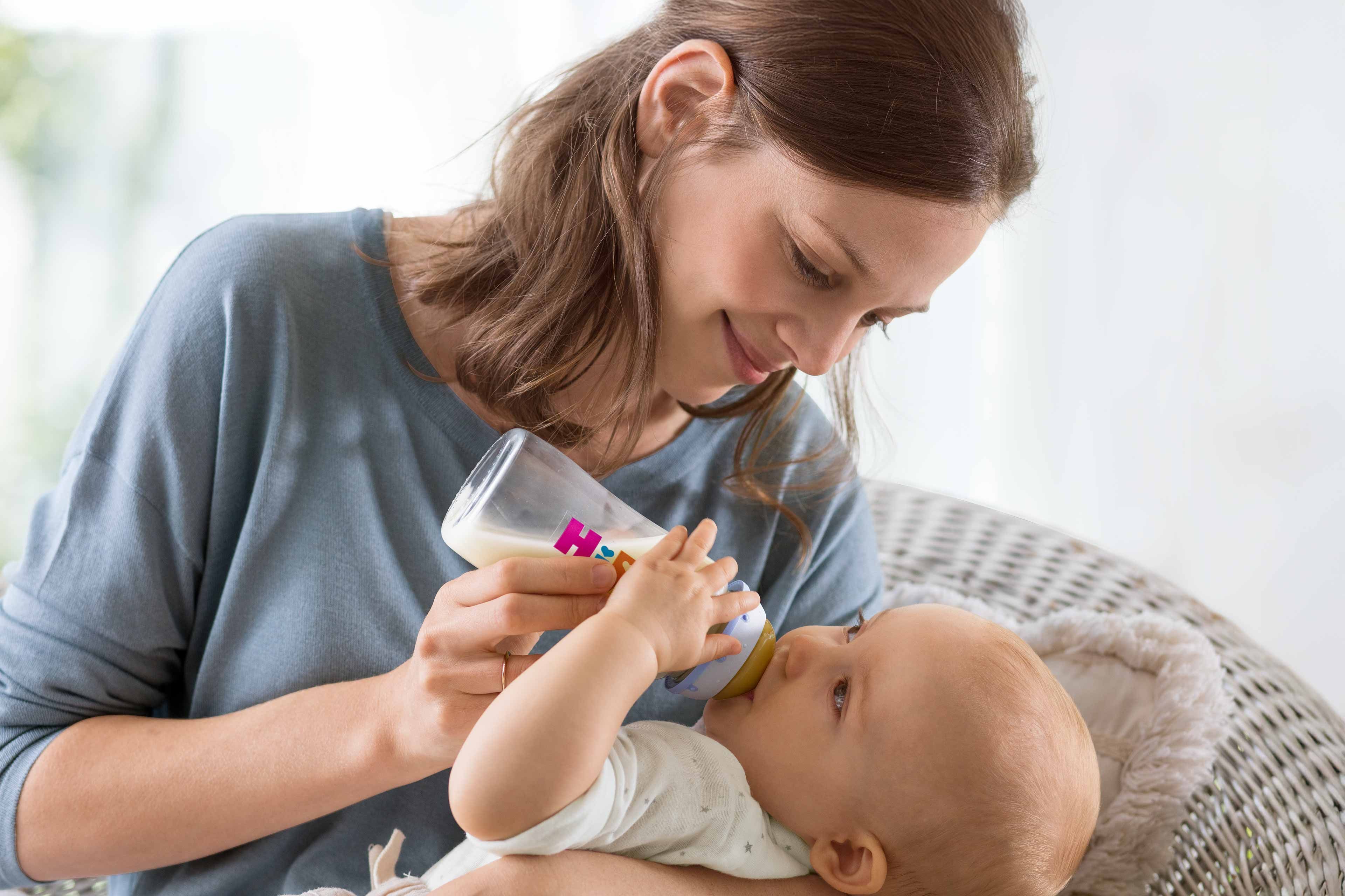 Eine Frau, die ihr Baby mit der Flasche füttert, während beide bequem in einem Raum mit Tageslicht sitzen.