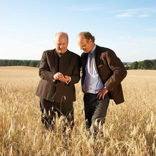 Zwei Männer im Anzug stehen in einem reifen Getreidefeld und begutachten Ähren vor weiter, naturbelassener Landschaft.