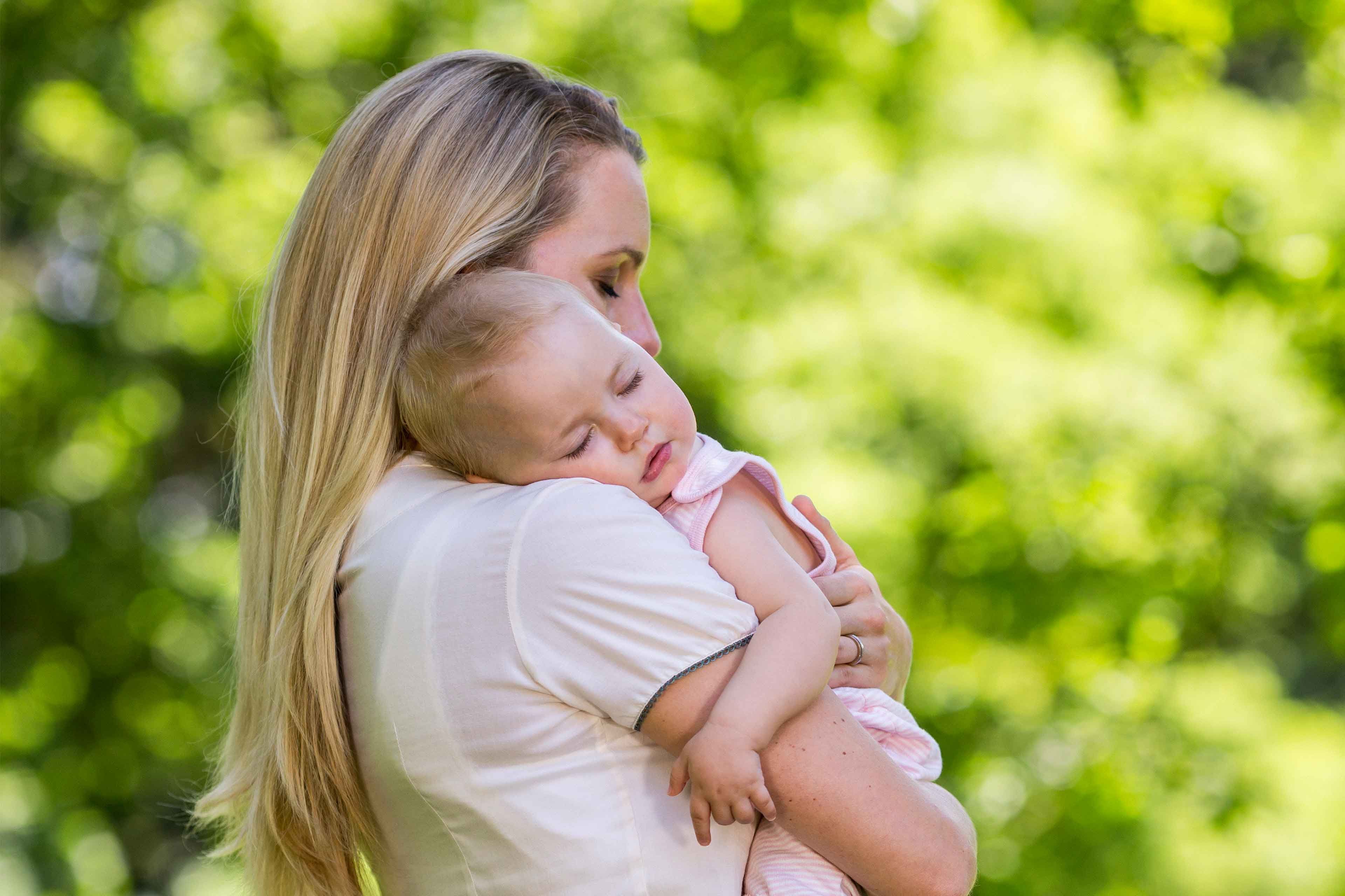 Mutter hält ihr schlafendes Baby liebevoll im Arm im Grünen, Symbol für Geborgenheit und ein entspanntes Babybäuchlein.