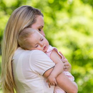 Mutter hält ihr schlafendes Baby liebevoll im Arm im Grünen, Symbol für Geborgenheit und ein entspanntes Babybäuchlein.