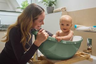 Lachende Mutter badet ihr fröhliches Baby in einer kleinen Wanne mit Schaum, umgeben von sanften Pflegeprodukten im Badezimmer.