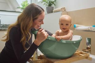 Lachende Mutter badet ihr fröhliches Baby in einer kleinen Wanne mit Schaum, umgeben von sanften Pflegeprodukten im Badezimmer.
