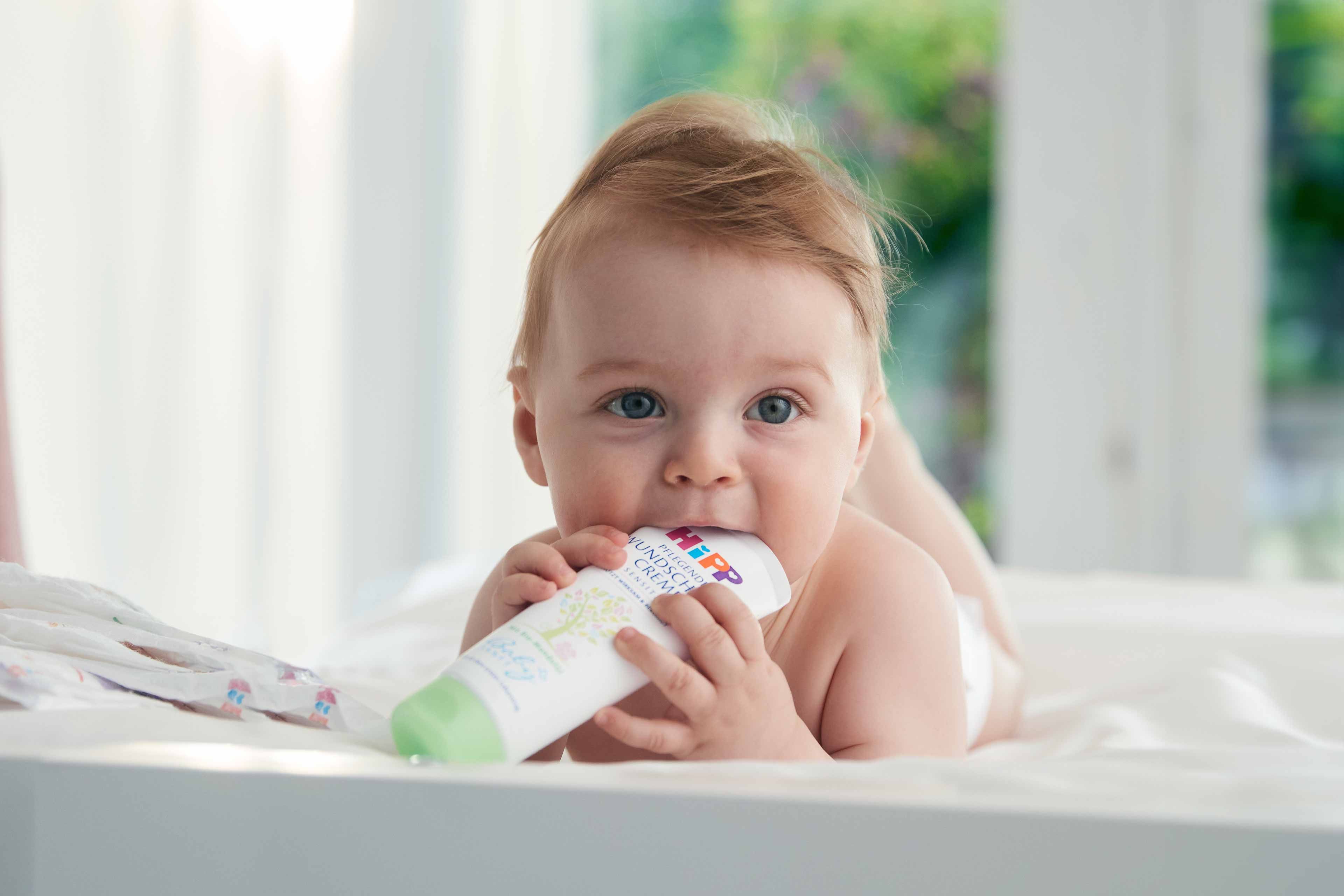 Ein Baby mit blauen Augen liegt auf einem Bett und kaut auf einer Tube Creme mit der Aufschrift "Wundschutz-Creme".