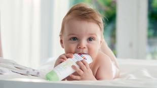 Ein Baby mit blauen Augen liegt auf einem Bett und kaut auf einer Tube Creme mit der Aufschrift "Wundschutz-Creme".