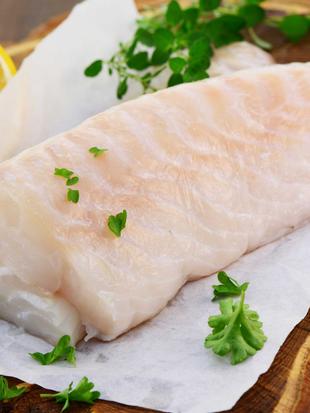 Ein Stück roher Fisch auf weißem Papier, garniert mit grünen Kräutern und einer Zitronenscheibe, auf einer Holzunterlage platziert.