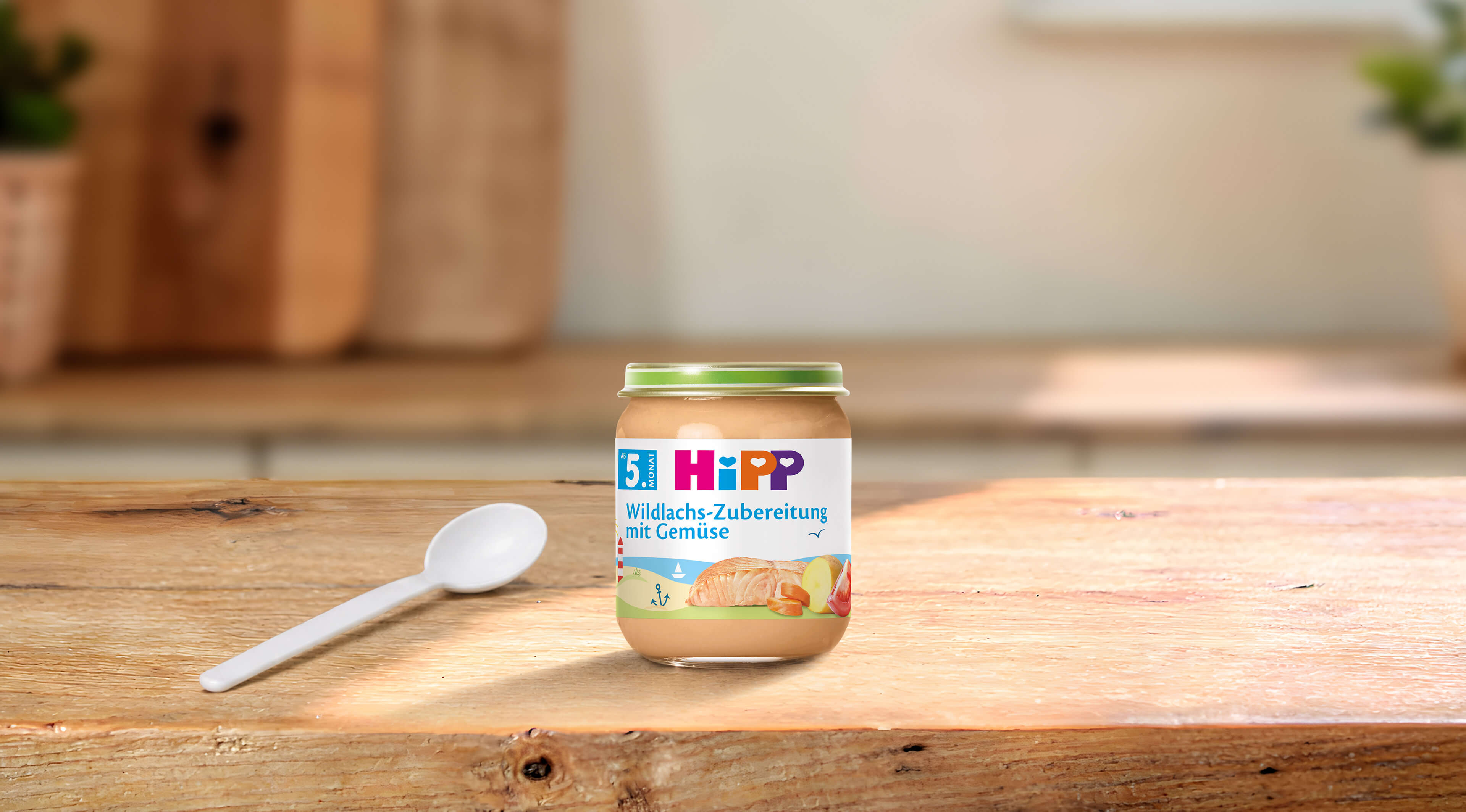 HiPP Wildlachs-Zubereitung mit Gemüse ab dem 5. Monat im Glas auf Holztisch, daneben ein weißer Babylöffel
