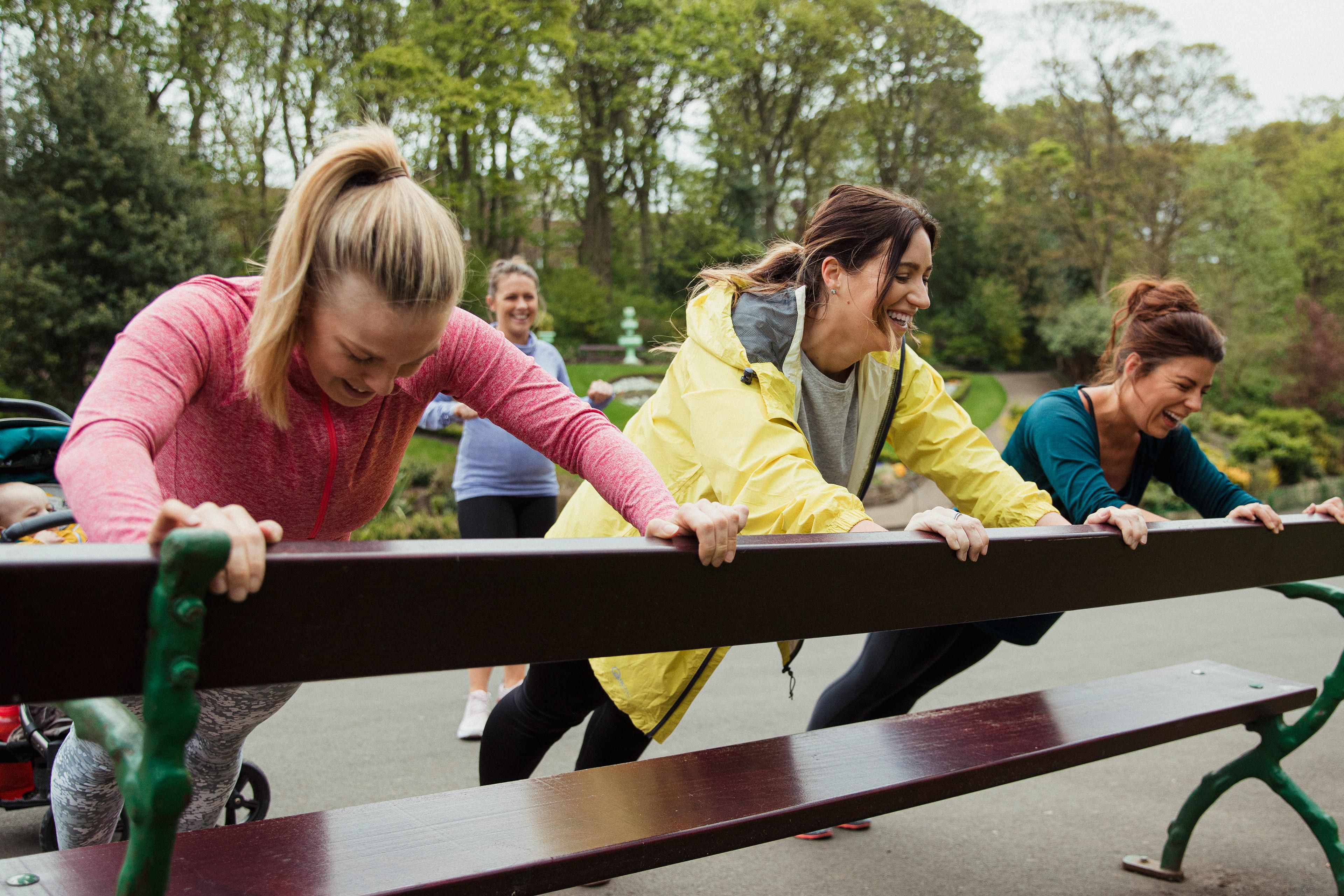Vier Frauen im Freien, die eine Fitnessaktivität ausüben, wobei eine von ihnen eine Bank schiebt.
