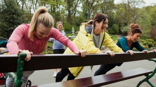 Vier Frauen im Freien, die eine Fitnessaktivität ausüben, wobei eine von ihnen eine Bank schiebt.