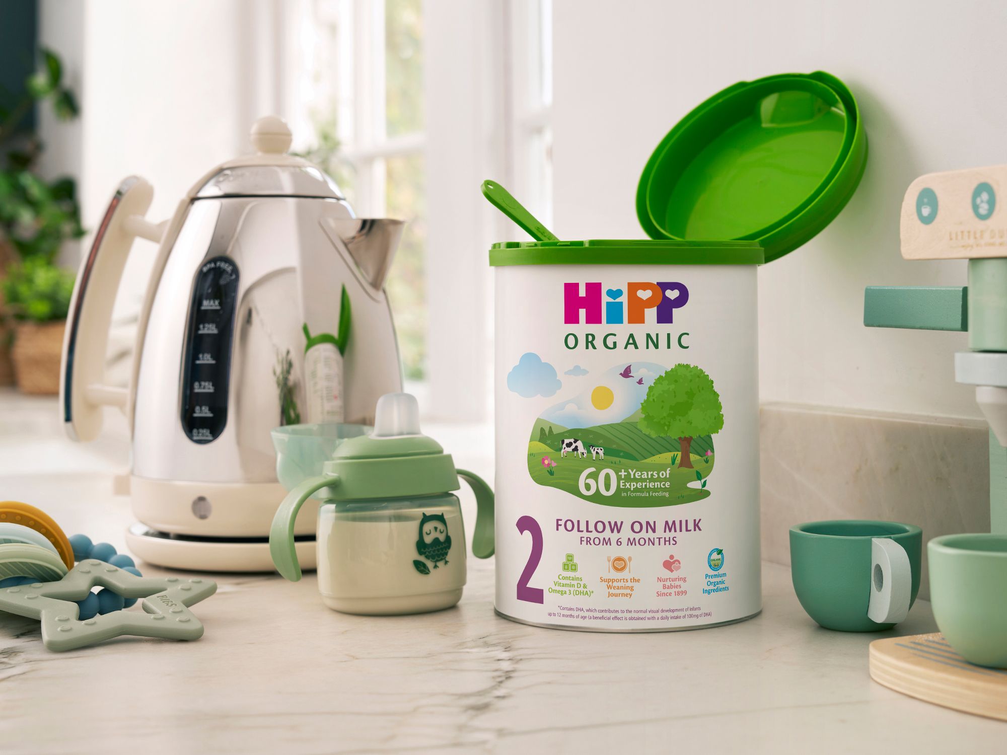 HiPP Organic Follow On Milk powder tin on a bright kitchen worktop beside a baby sippy cup, kettle and baby toys.