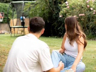 Couple relaxing on the grass watching their child play on a trampoline in a leafy garden