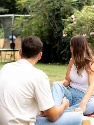 Couple relaxing on the grass watching their child play on a trampoline in a leafy garden
