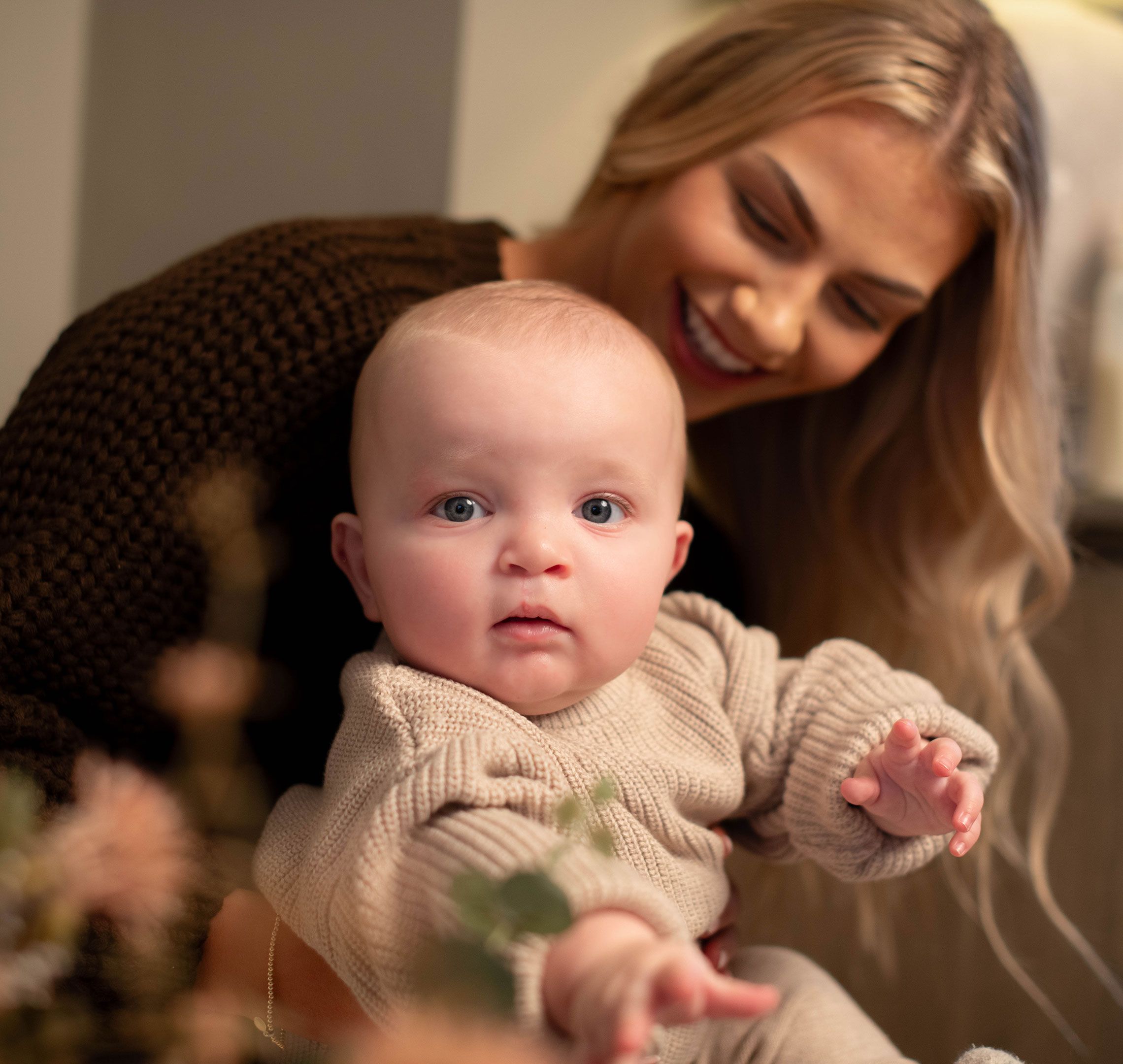 Baby in a cosy knitted outfit reaching forward while being gently held by a smiling parent in a warm, calm home setting.
