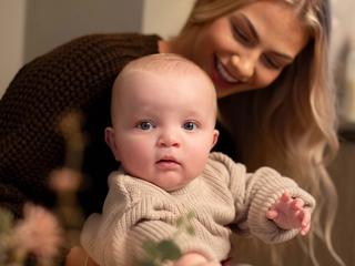 Baby in a cosy knitted outfit reaching forward while being gently held by a smiling parent in a warm, calm home setting.