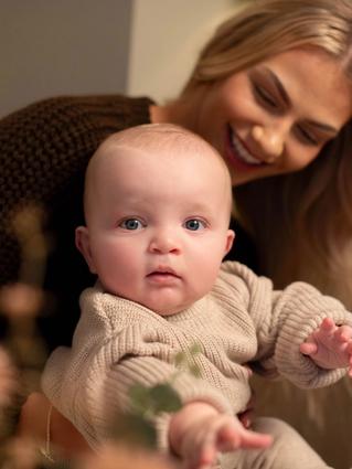 Baby in a cosy knitted outfit reaching forward while being gently held by a smiling parent in a warm, calm home setting.