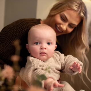 Baby in a cosy knitted outfit reaching forward while being gently held by a smiling parent in a warm, calm home setting.