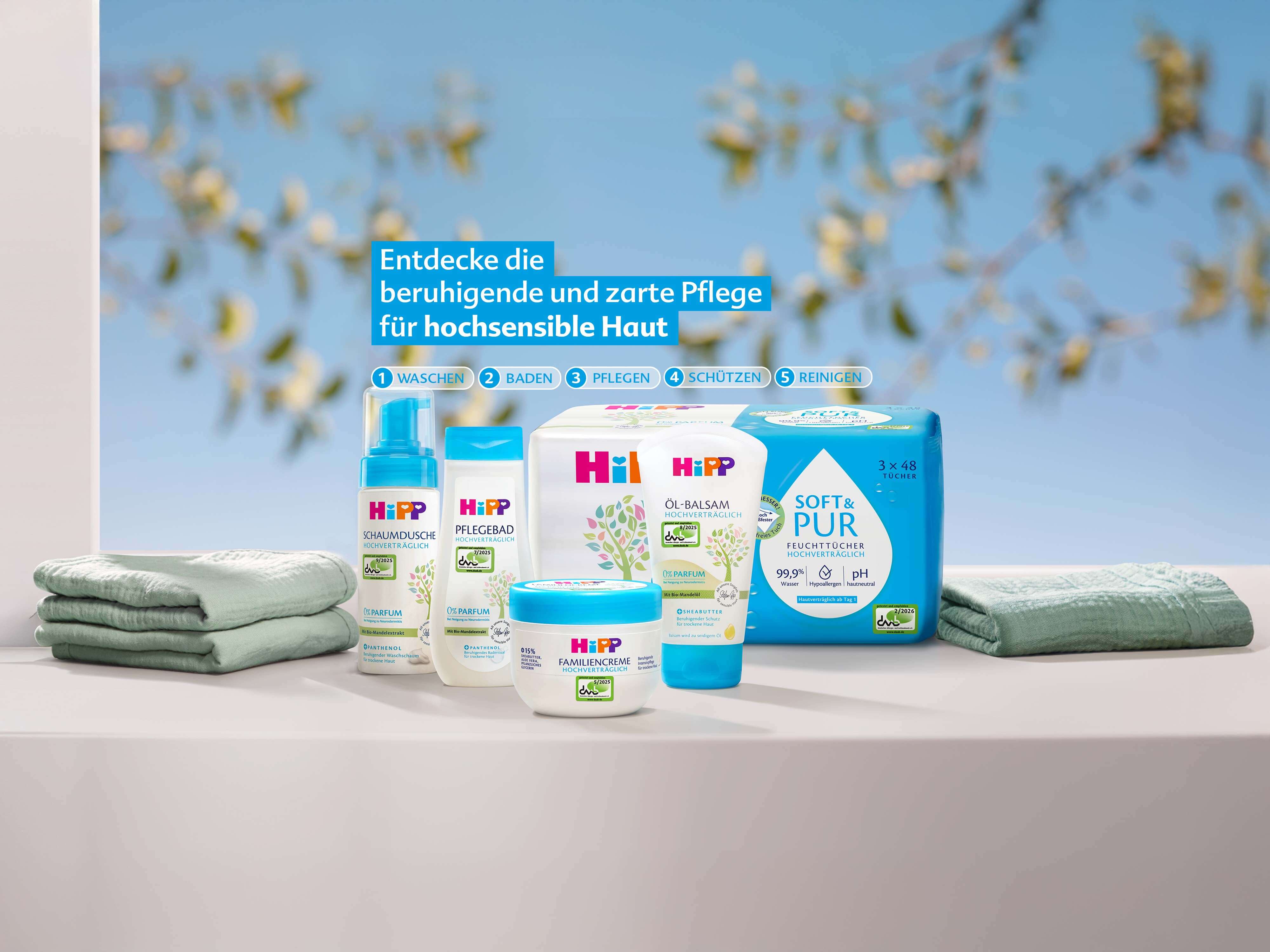 Mehrere HiPP-Pflegeprodukte für sensible Haut stehen auf einem Tisch, umgeben von gefalteten Handtüchern und einem unscharfen Hintergrund mit gelben Blüten.