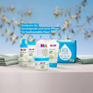 Mehrere HiPP-Pflegeprodukte für sensible Haut stehen auf einem Tisch, umgeben von gefalteten Handtüchern und einem unscharfen Hintergrund mit gelben Blüten.
