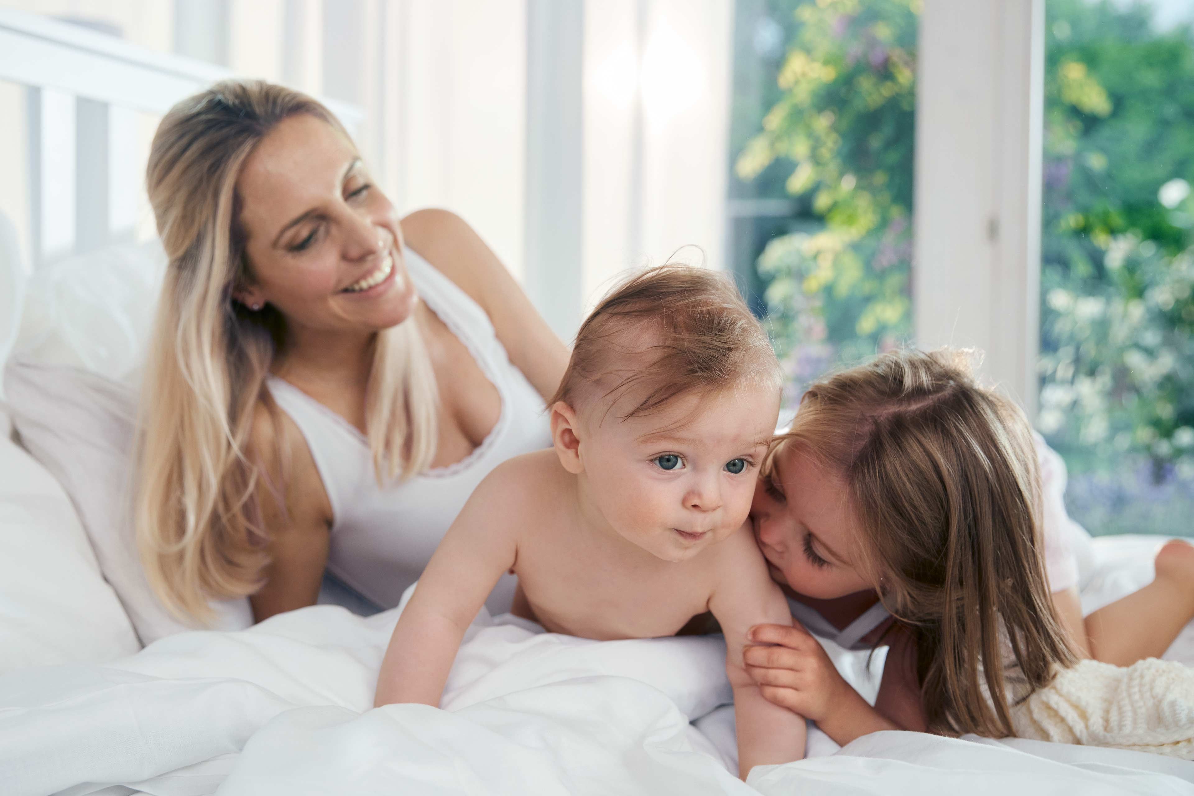 Ein fröhlicher Moment zwischen einer Mutter und ihren beiden Kindern, wobei die Mutter lächelt und das Baby neugierig in die Kamera schaut.