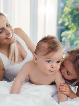 Ein fröhlicher Moment zwischen einer Mutter und ihren beiden Kindern, wobei die Mutter lächelt und das Baby neugierig in die Kamera schaut.