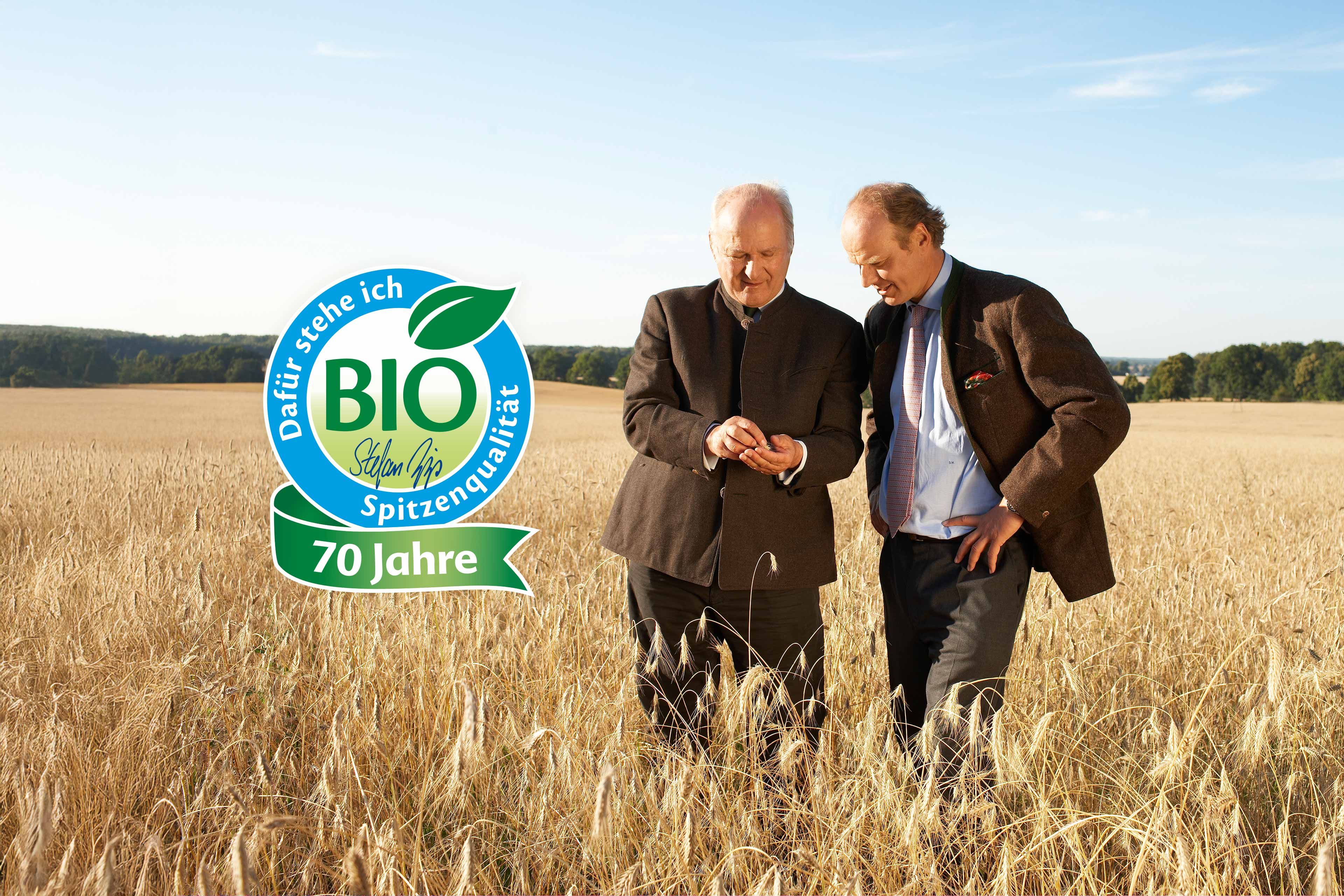 Zwei Männer begutachten Getreide auf einem Bio-Feld, daneben ein Siegel „BIO Spitzenqualität – 70 Jahre“ vor weiter Naturkulisse.