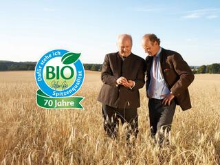 Zwei Männer begutachten Getreide auf einem Bio-Feld, daneben ein Siegel „BIO Spitzenqualität – 70 Jahre“ vor weiter Naturkulisse.