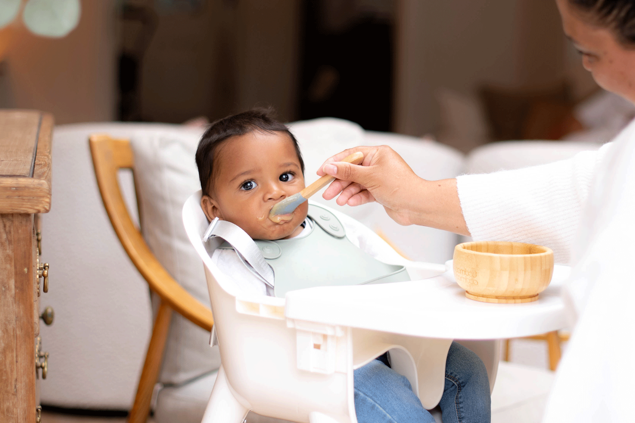 A dark-haired baby in blue jeans sits in a white high chair wearing a sage-green silicone bib, looking straight ahead as a mother feeds them purée with a wooden spoon. On the white tray sits a small wooden suction bowl, with a soft-focus living room background featuring a wooden cabinet and white sofa cushions.