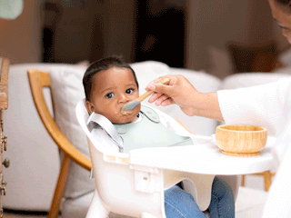 A dark-haired baby in blue jeans sits in a white high chair wearing a sage-green silicone bib, looking straight ahead as a mother feeds them purée with a wooden spoon. On the white tray sits a small wooden suction bowl, with a soft-focus living room background featuring a wooden cabinet and white sofa cushions.