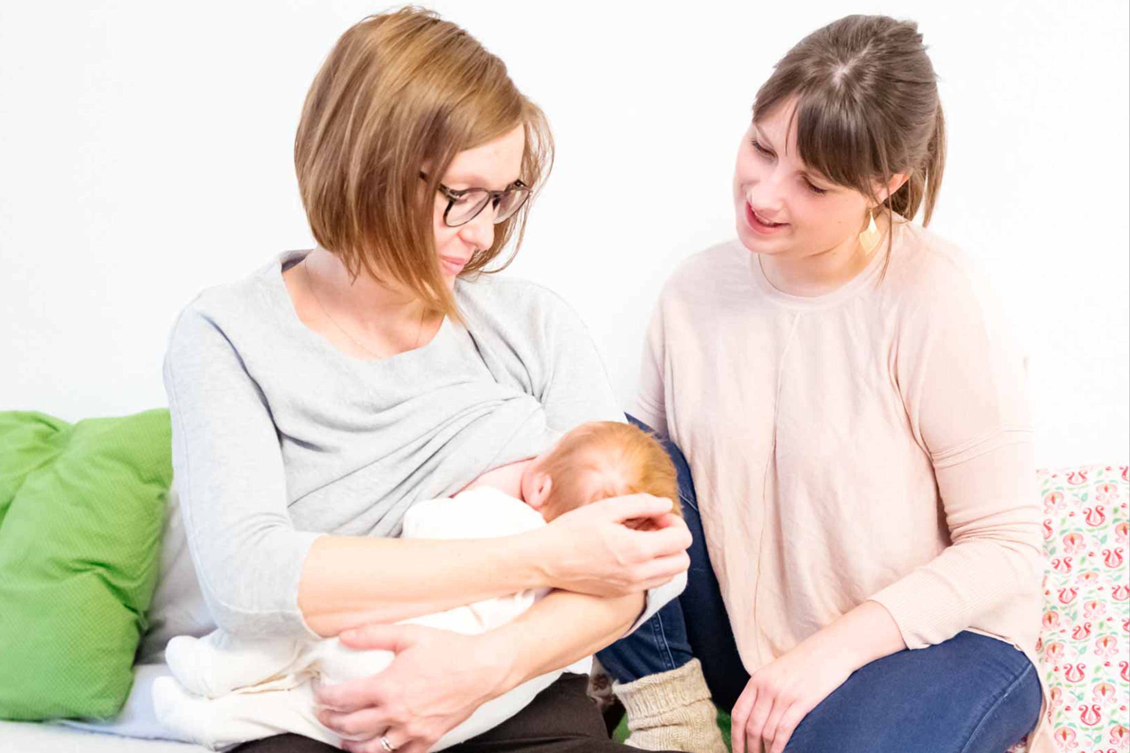 Zwei Frauen, eine davon mit einem Baby in der Hand, in einem Raum mit weißem Hintergrund, mit einem grünen Kissen und einem blumengemusterten Kissen im Hintergrund.