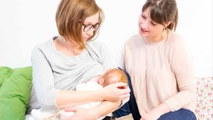 Zwei Frauen, eine davon mit einem Baby in der Hand, in einem Raum mit weißem Hintergrund, mit einem grünen Kissen und einem blumengemusterten Kissen im Hintergrund.