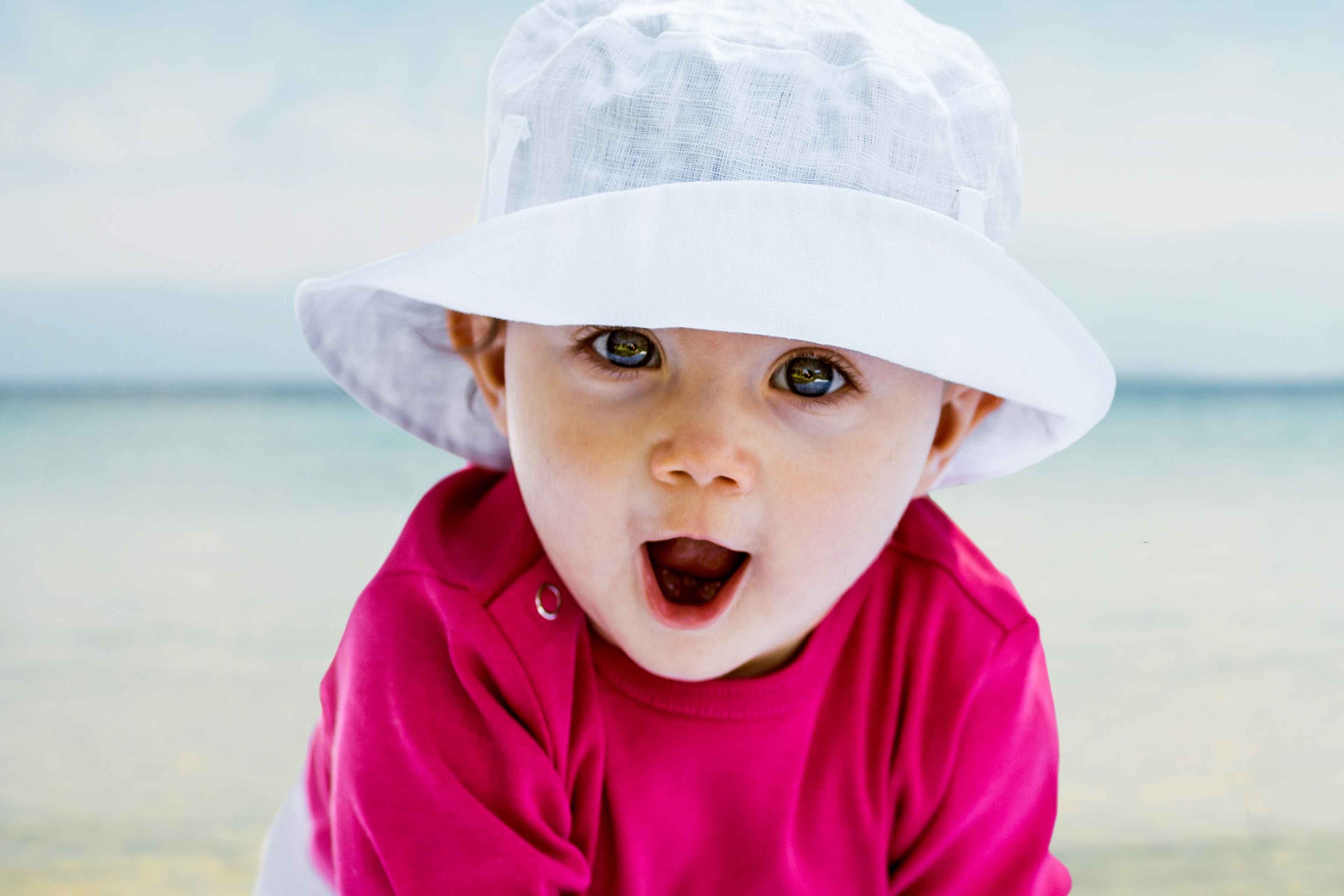 Ein kleines Kind mit leuchtend blauen Augen, das eine weiße Mütze und ein rosa Hemd trägt, mit offenem Mund, als ob es überrascht wäre.