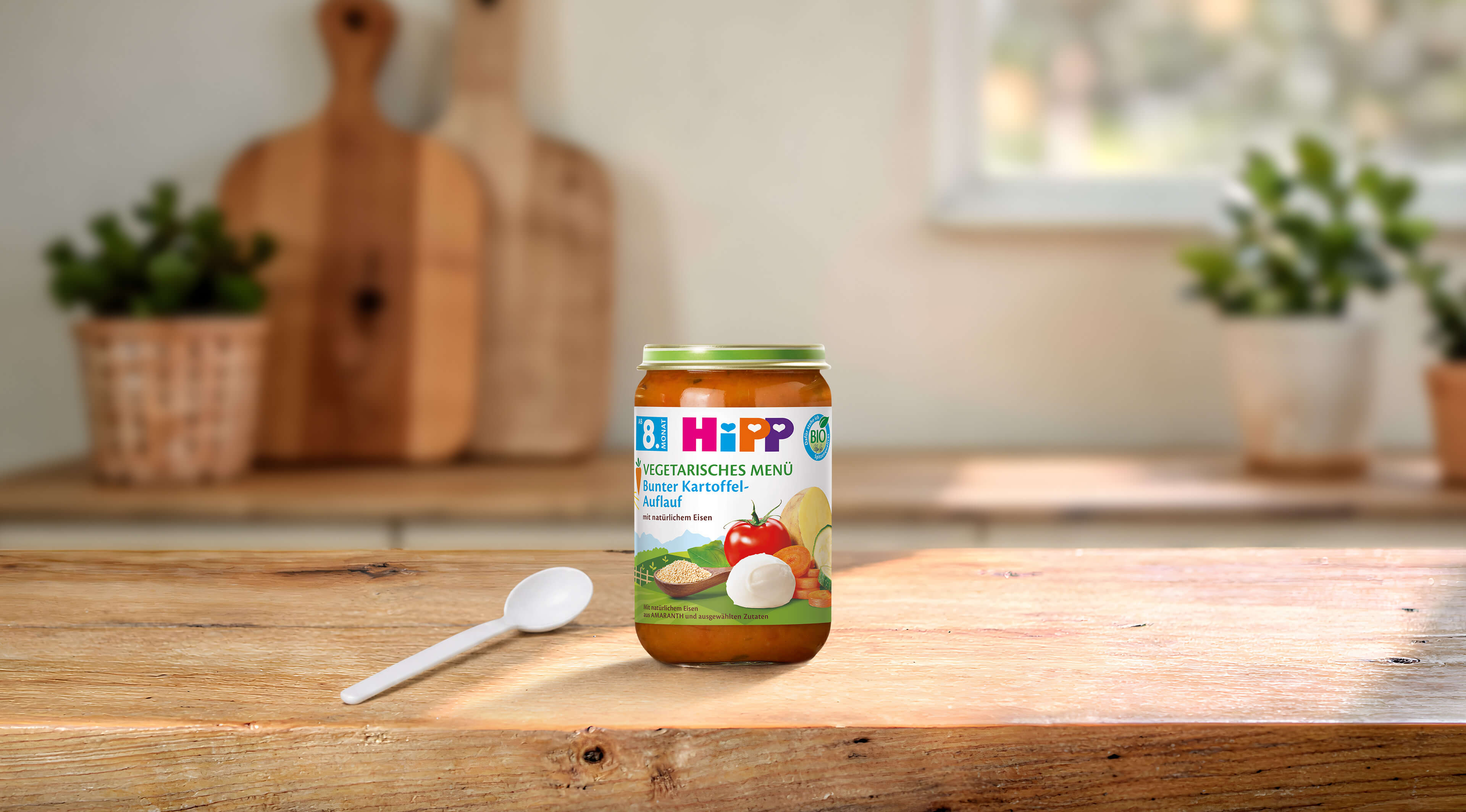 HiPP Bio Vegetarisches Menü Bunter Kartoffel-Auflauf im Glas mit Löffel auf Holztisch in heller Familienküche