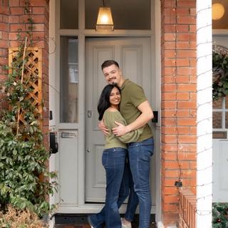 Couple hugging and smiling in front of their home decorated with Christmas lights and greenery.