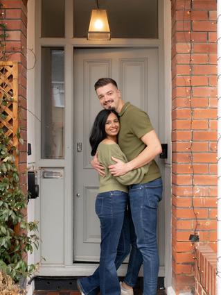 Couple hugging and smiling in front of their home decorated with Christmas lights and greenery.