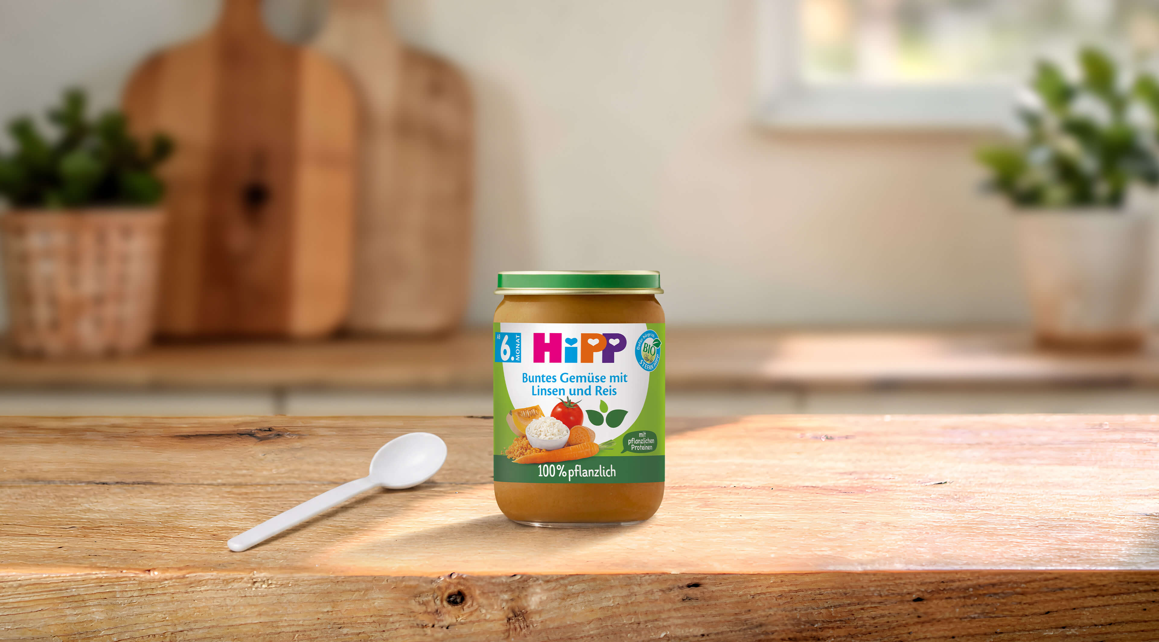 HiPP Bio-Gläschen „Buntes Gemüse mit Linsen und Reis“ ab 6. Monat auf Holztisch mit Löffel in heller Küche