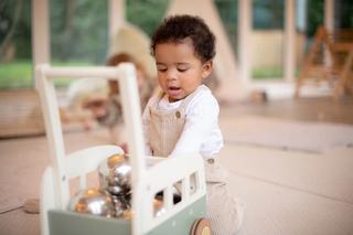 A toddler wearing beige overalls and a white shirt plays with a wooden pushcart filled with shiny silver balls. The child is focused and engaged in a bright indoor play area with soft flooring and a tent in the background.