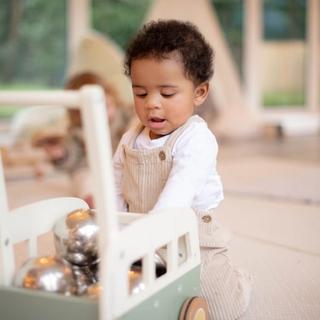 A toddler wearing beige overalls and a white shirt plays with a wooden pushcart filled with shiny silver balls. The child is focused and engaged in a bright indoor play area with soft flooring and a tent in the background.