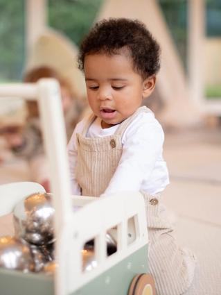 A toddler wearing beige overalls and a white shirt plays with a wooden pushcart filled with shiny silver balls. The child is focused and engaged in a bright indoor play area with soft flooring and a tent in the background.
