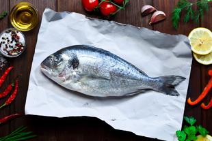 Ein frischer Fisch auf weißem Papier, umgeben von verschiedenen Zutaten wie Chilischoten, Zitrone, Knoblauch und Kräutern, alles vor einem Holzhintergrund.