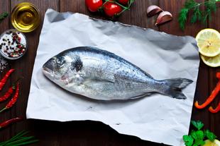Ein frischer Fisch auf weißem Papier, umgeben von verschiedenen Zutaten wie Chilischoten, Zitrone, Knoblauch und Kräutern, alles vor einem Holzhintergrund.