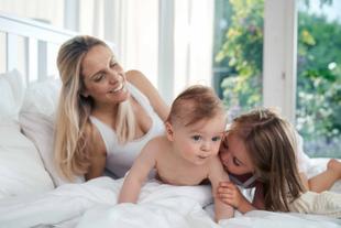 Ein fröhlicher Moment zwischen einer Mutter und ihren beiden Kindern, wobei die Mutter lächelt und das Baby neugierig in die Kamera schaut.