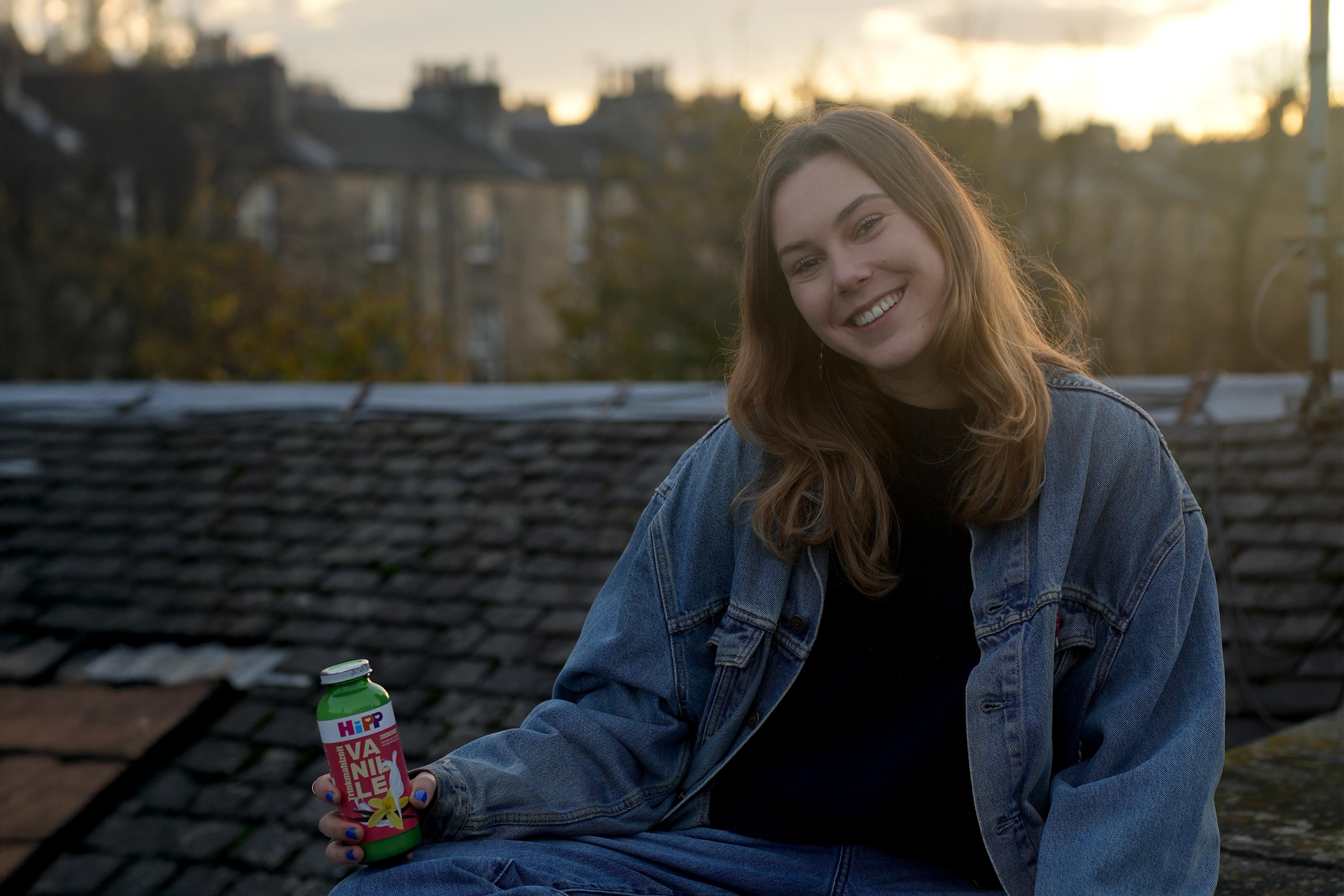 Person mit Jeansjacke sitzt auf einem Dach und hält eine Flasche HiPP Vanille Trinkmahlzeit