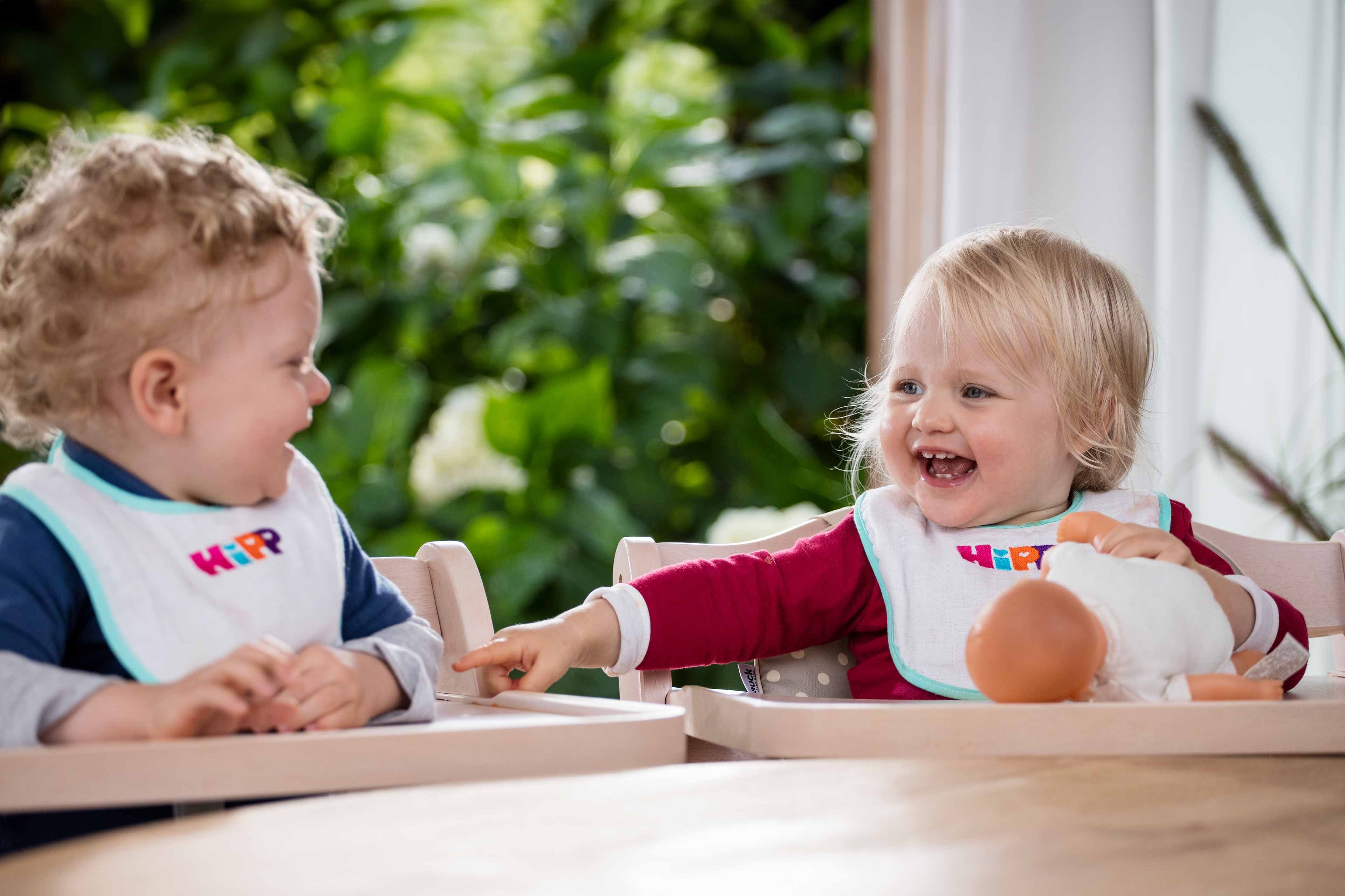 Zwei Kleinkinder, eines in einem blauen Hemd und das andere in einem roten Outfit, sitzen in Hochstühlen und spielen mit einer Puppe.