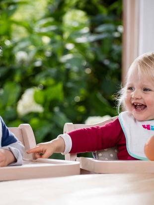 Zwei Kleinkinder, eines in einem blauen Hemd und das andere in einem roten Outfit, sitzen in Hochstühlen und spielen mit einer Puppe.