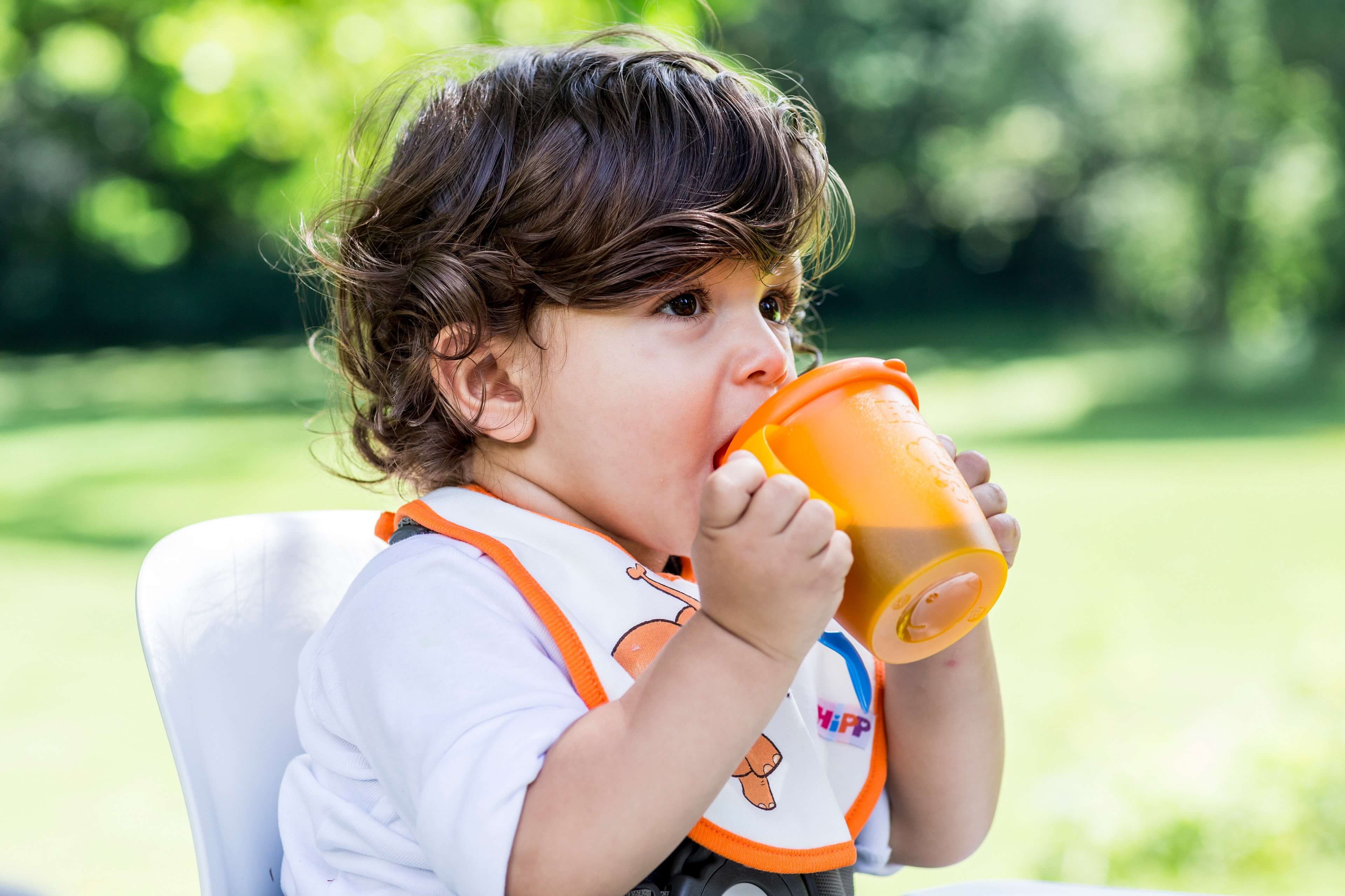 Ein kleines Kind, vermutlich ein Kleinkind, nippt im Freien an einer orangefarbenen Schnabeltasse.
