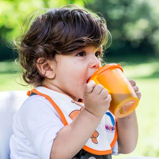 Ein kleines Kind, vermutlich ein Kleinkind, nippt im Freien an einer orangefarbenen Schnabeltasse.