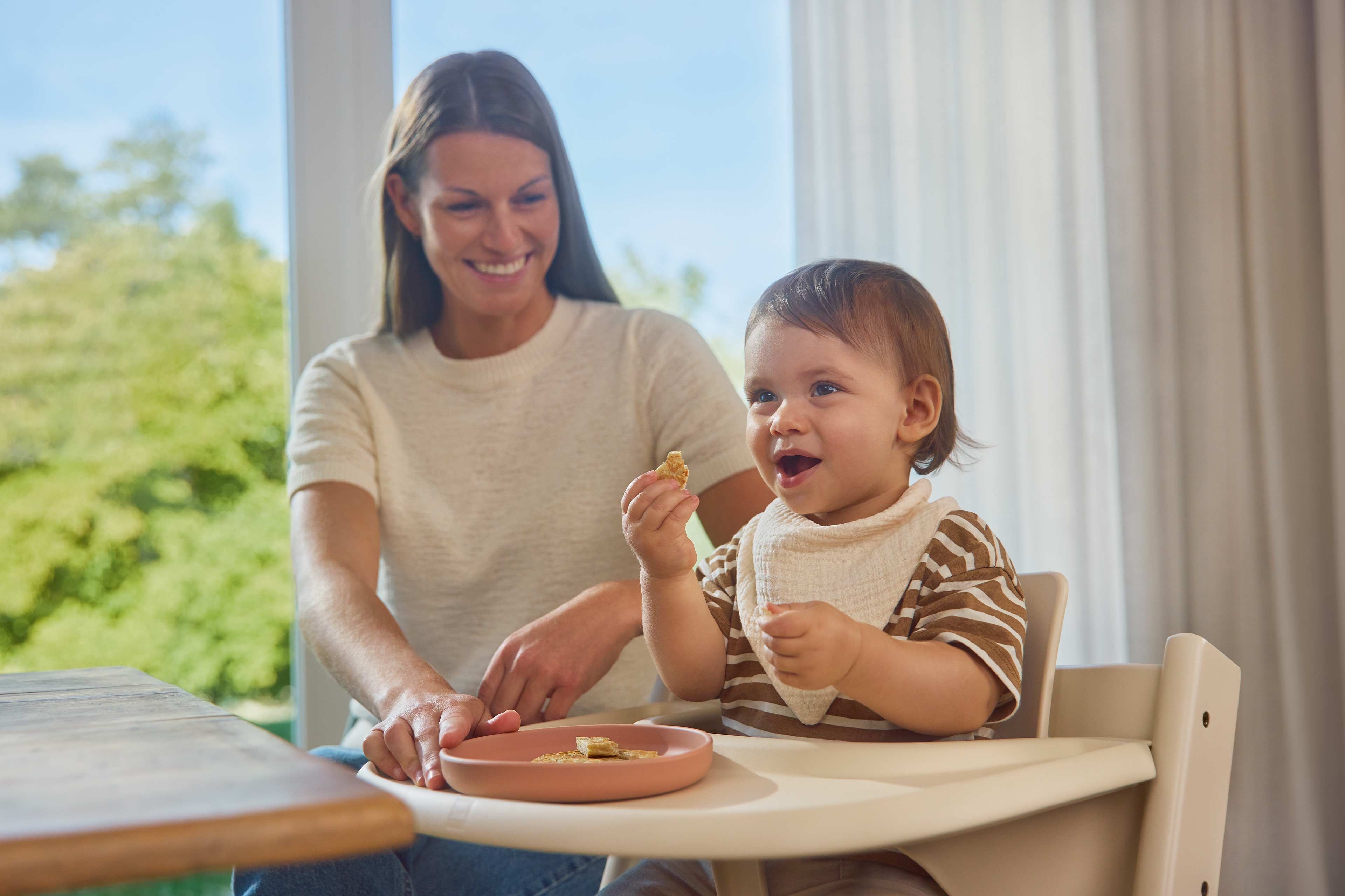 Fröhliches Baby isst beim Baby Led Weaning selbstständig Fingerfood, während seine Mutter es begleitet.