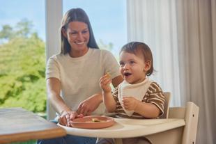 Lächelnde Mutter begleitet ihr Kleinkind beim selbstständigen Essen im Hochstuhl.