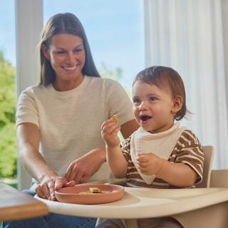 Fröhliches Baby isst beim Baby Led Weaning selbstständig Fingerfood, während seine Mutter es begleitet.