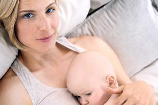 Eine Frau mit blondem Haar und blauen Augen, die ein Baby an sich drückt, das auf einer weichen Unterlage liegt.